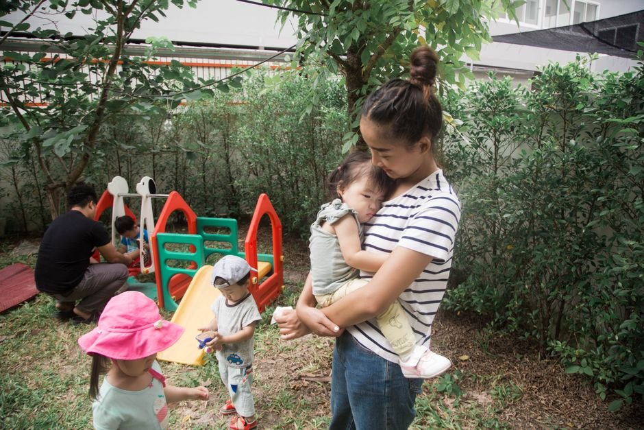Mama glėbyje laiko kūdikį, kiti vaikai džiaugsmingai žaidžia kiemo žaidimų aikštelėje
