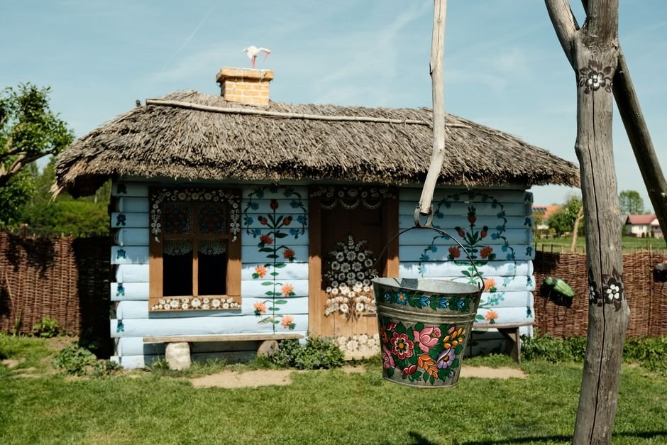 Kaimiškas namelis su gėlėtais piešiniais, ryškiu kibirėliu ir šiaudiniu stogu – žaismingas vaikų kampelis